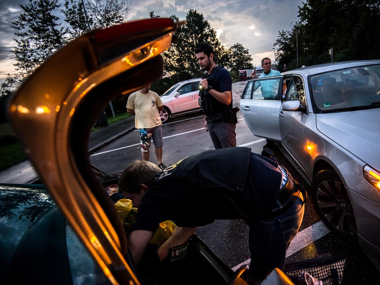 Ein Bundespolizist durchsucht den Kofferraum eines Autos, im Hintergrund stehen mehrere Männer herum.