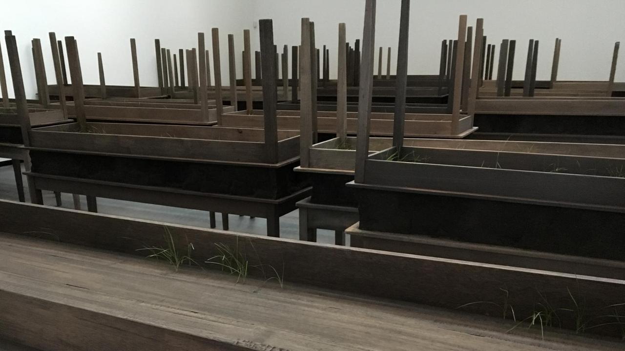 Die Werkgruppe "Plegaria muda" der Künstlerin Doris Salceda: Gestapelte Tische, durch deren Holz Gras wächst.