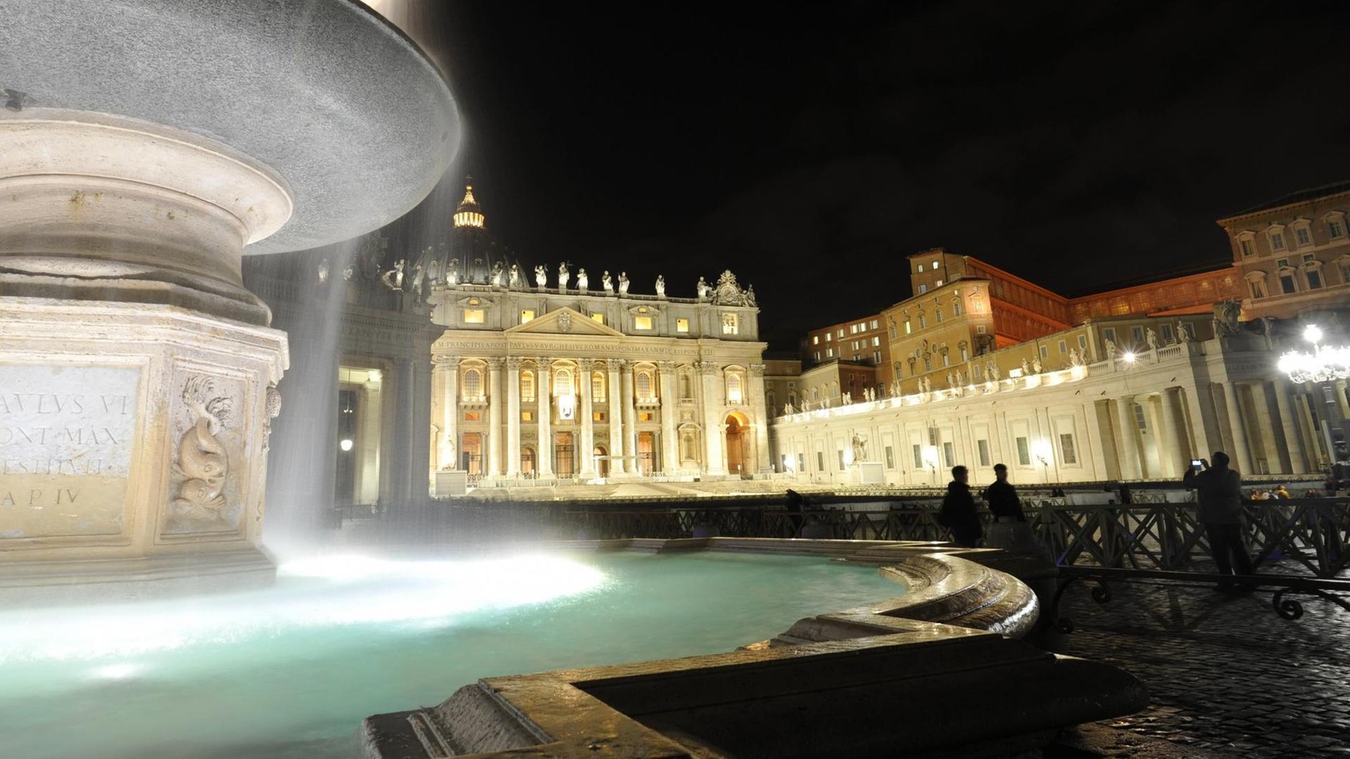 Ein Brunnen auf dem Petersplatz wird beleuchtet, im Hintergrund steht der Petersdom.