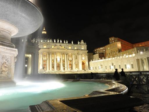 Ein Brunnen auf dem Petersplatz wird beleuchtet, im Hintergrund steht der Petersdom.