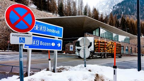 Ein mit Baumstämmen beladener Lkw befährt den Brennerpass. Im Hintergrund die schneebedeckten Alpen und Tannenwälder, im Vordergrund sieht man zwei Schilder Richtung Innsbruck und Bozen.