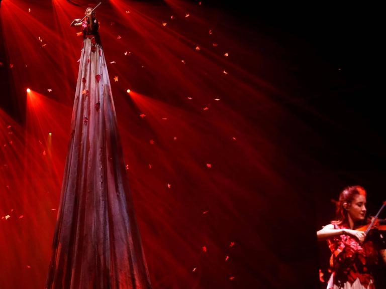 Szene aus der Show "Game of Thrones Live Experience", welche die Serienmusik live präsentiert: eine Frau spielt vor rotem Licht mit Glitzerelementen auf Stelzen ein Streichinstrument, eine andere spielt im Vordergrund ein Streichinstrument.
