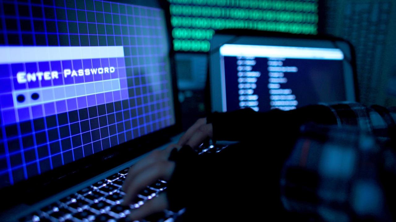 Ein Passwort wird auf einem Laptop über die Tastatur eingegeben. Die Hände auf der Tastatur tragen schwarze Stulpen. Auf dem Monitor sind die Worte "Enter Password" zu lesen. Im Hintergrund erkennt man verschwommen weitere Bildschirme. 