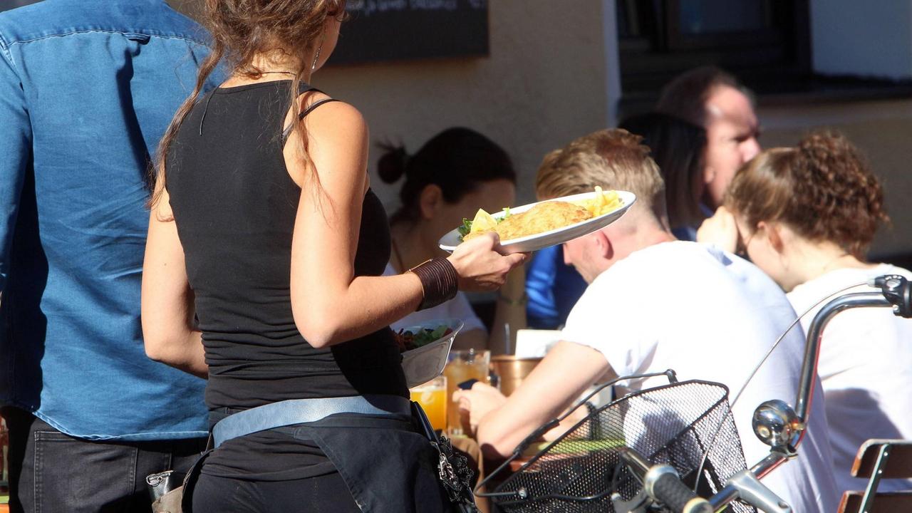 Eine Kellnerin trägt Speisen und Getränke an einem Tisch im Außenbereich eines Cafés in München bei strahlendem Sonnenschein.