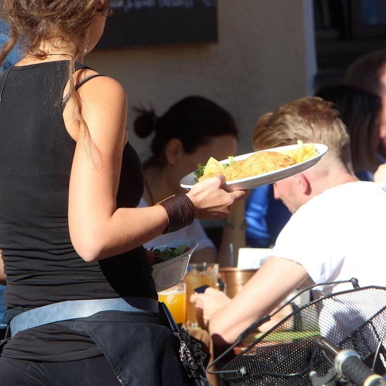 Eine Kellnerin trägt Speisen und Getränke an einem Tisch im Außenbereich eines Café s in München bei strahlendem Sonnenschein.