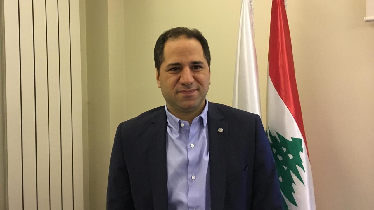 Samy Gemayel ist Abgeordneter im Libanon - schlank, dunkle Haare, Sakko präsentiert er sich vor Libanons Flagge.