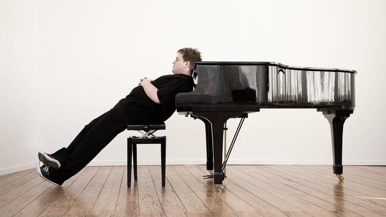 Matthias Reuter sitzt auf einem Klavierhocker und lehnt sich mit dem Rücken gegen einen schwarzen Flügel.