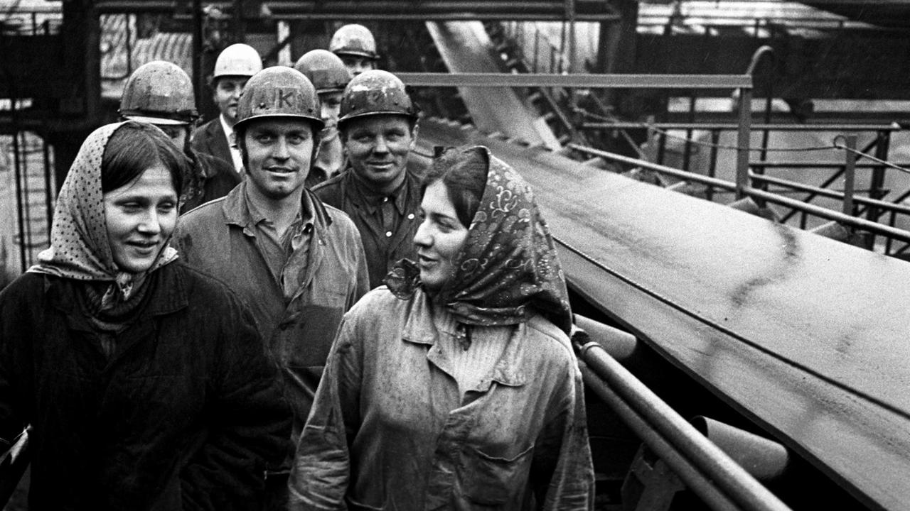 Arbeiter und Arbeiterinnen stehen neben einem Förderband auf dem Betriebsgelände des VEB Gaskombinat Schwarze Pumpe.