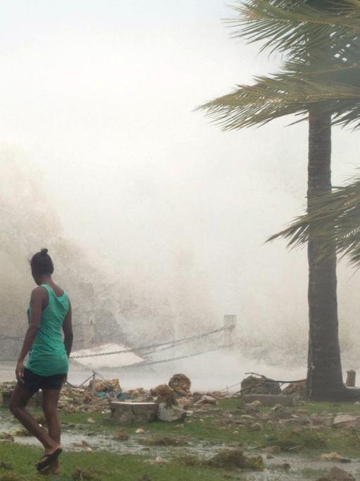 Eine Frau geht vor einer aufbrausenden Welle am Strand entlang. Palmen biegen sich im Sturm. Am Strand liegen Trümmer.