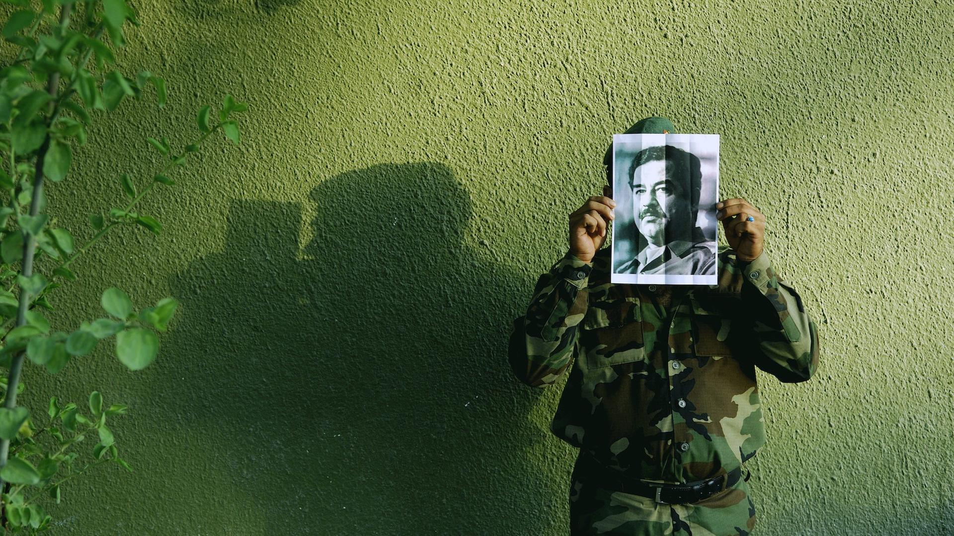 Ein Soldat vor einer grünen Hauswand hält ein fotokopiertes Porträt von Saddam Hussein vor sein Gesicht.
