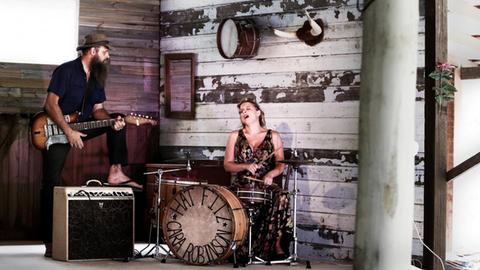 Eine Frau im geblümten Kleid sitzt an einem Schlagzeug. Ein Mann mit Hut hält eine Gitarre in der Hand. Er stützt ein Bein auf einem Lautsprecher ab. Sie befinden sich in einer Scheune.