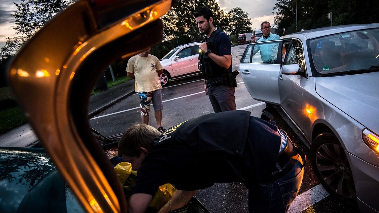 Ein Bundespolizist durchsucht den Kofferraum eines Autos, im Hintergrund stehen mehrere Männer herum.