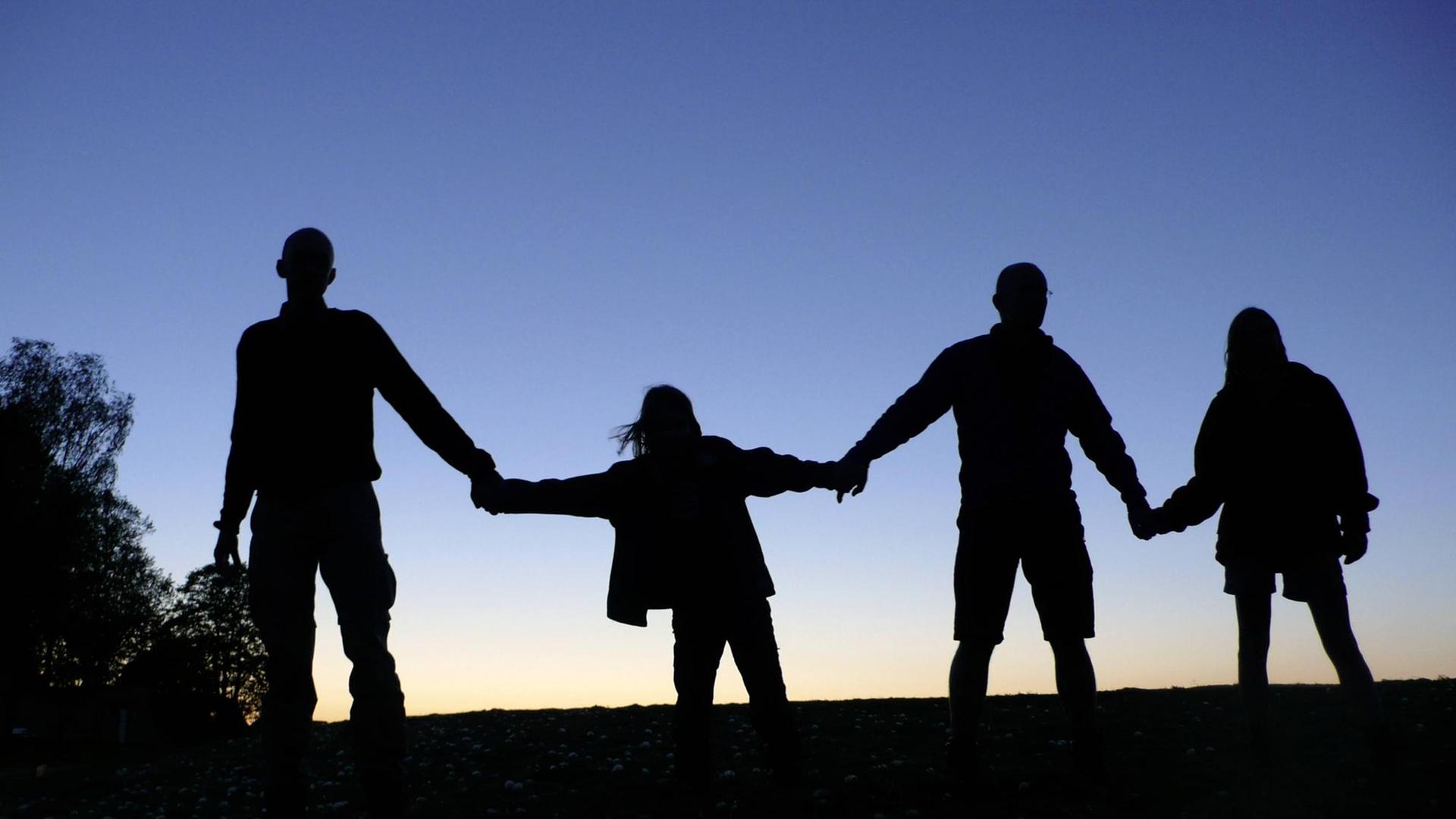 Drei Erwachsene und ein Kind halten sich die Hände, sie sind als Schattenriss vor dem dämmernden Himmel zu sehen. (Symbolbild gesellschaftlicher Zusammenhalt)