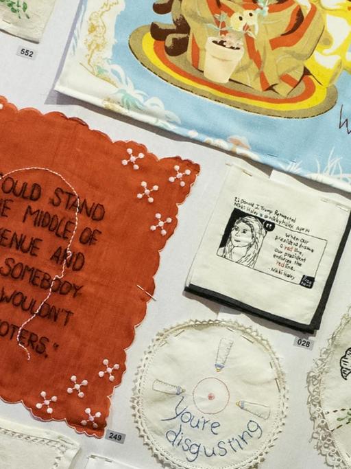 Alte Textilien liegen ausgebreitet nebeneinander, auf die die Künstlerin Diane Weymar verstörende Aussagen von Präsident Donald Trump gestickt hat. "Tiny Pricks", auf deutsch "Kleine Stiche", heißt die Arbeit.