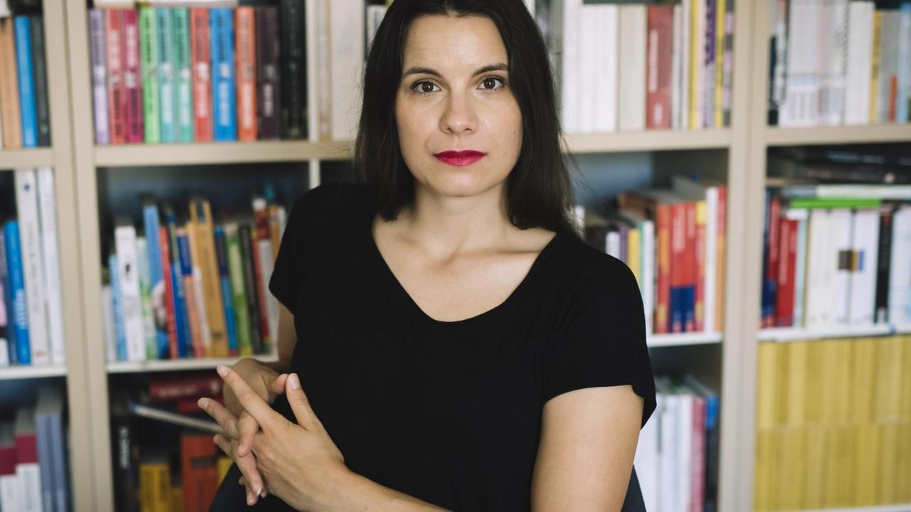 Die Autorin Didi Drobna sitzt vor einem Bücherregal und guckt selbstbewusst in die Kamera