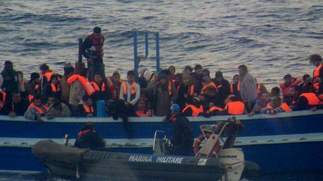 Flüchtlinge auf einem Boot, davor ein Rettungsboot der italienischen Marine.