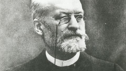 Eine schwarz-weiße Fotografie zeigt Er war den französischen Mediziner und Bakteriologen Charles Louis Alphonse Laveran. Er trägt einen graue Vollbart und randlose Brille.