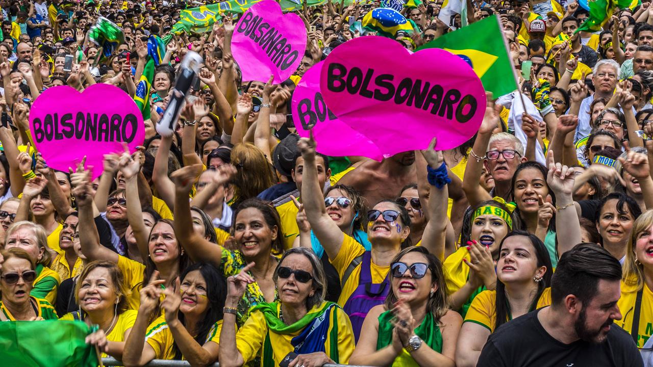 Eine Masse von Leuten in brasilianischen Nationalfarben applaudieren und halten rosafarbene, herzförmige Schilder mit Bolsonaros Namen hoch.