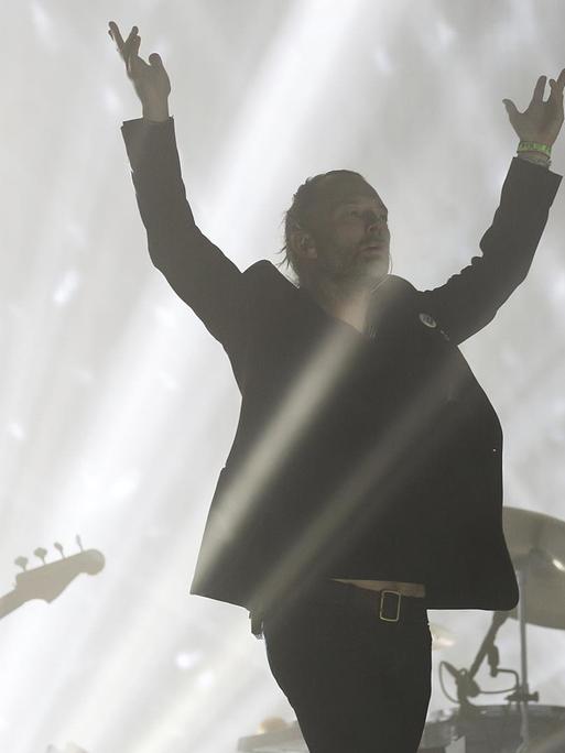 Ein Mann steht vor einem Schlagzeug auf einer Bühne im Laserlicht und hebt die Arme. Hinter spielt jemand E-Bass.