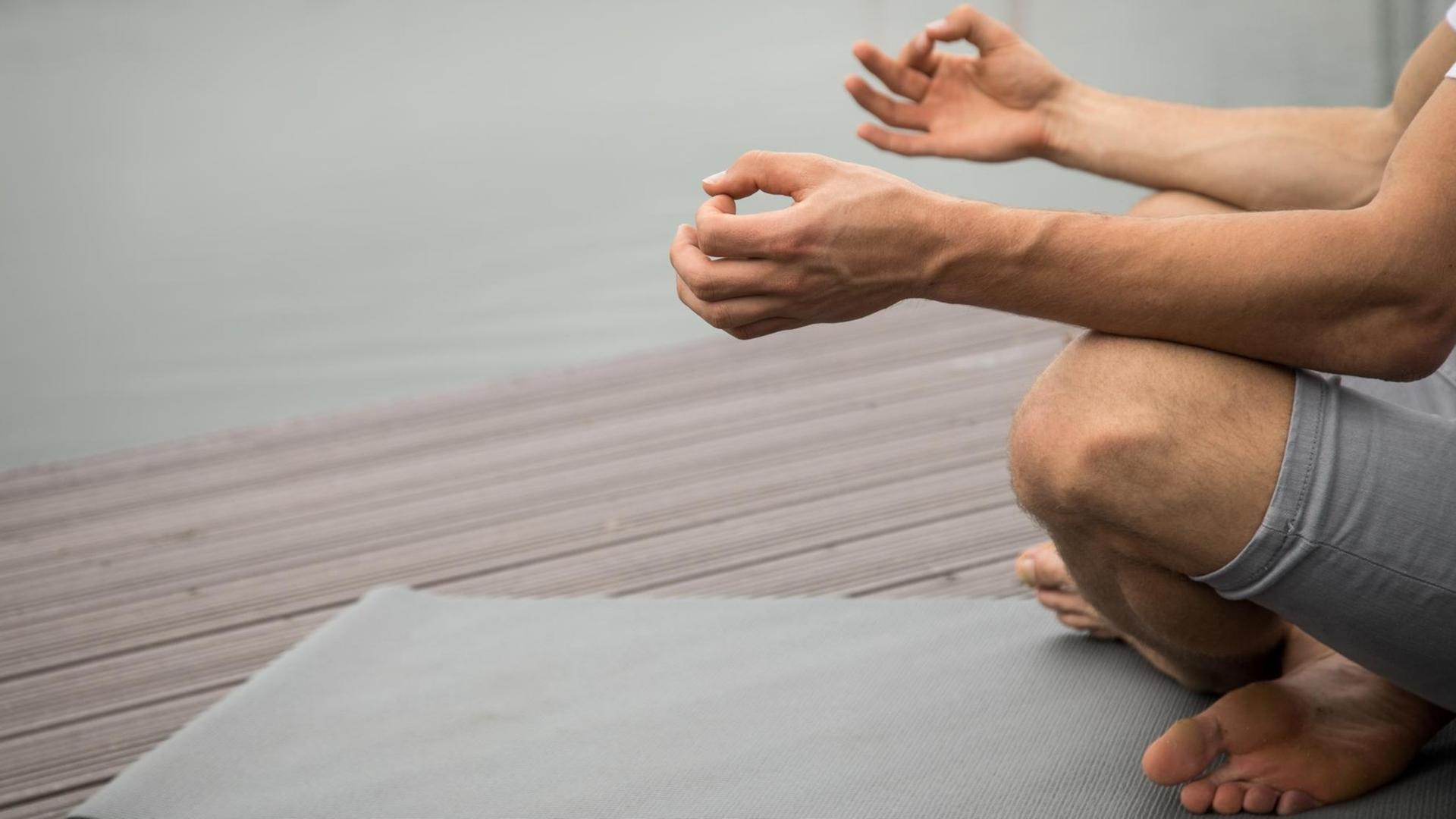 Ein Mann sitzt an einem Seeufer und meditiert auf einer Yogamatte