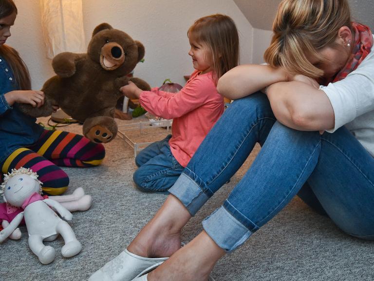 Gestresst sitzt eine Mutter im Kinderzimmer, während sich dahinter ihre beiden Kinder um ein Plüschtier streiten.
