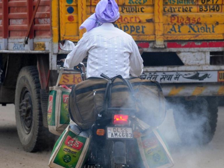 Auf dem Motorrad sitzt ein Mann mit lila Turban. Das Motorrad stößt weißen rauch aus. Es fährt hinter einem orangefarbenen LKW her.