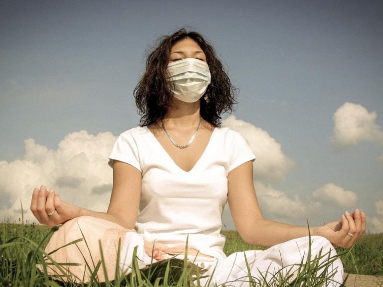 Eine Frau mit Mundschutz sitzt auf einer Wiese und meditiert.