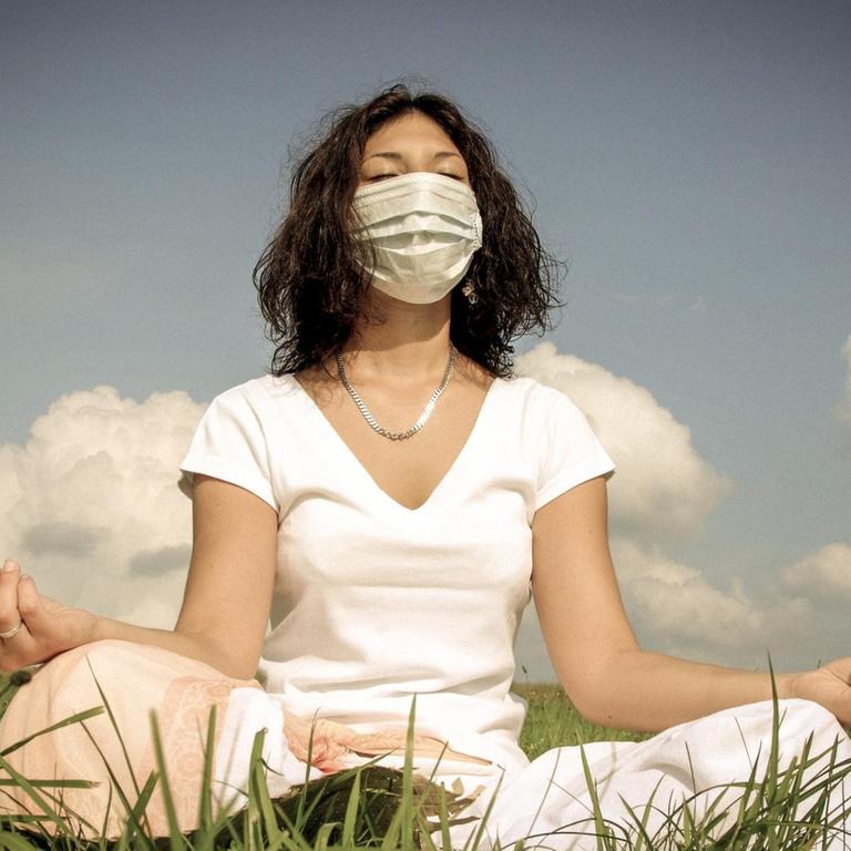Eine Frau mit Mundschutz sitzt auf einer Wiese und meditiert.