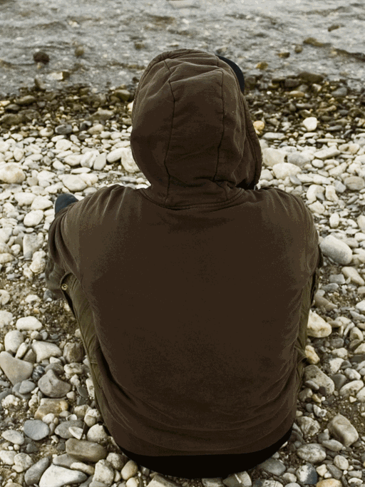 Ein junger Mann sitzt mit dem Rücken zur Kamera an einem Flußufer. Er trägt eine Shirt mit einer Kapuze.