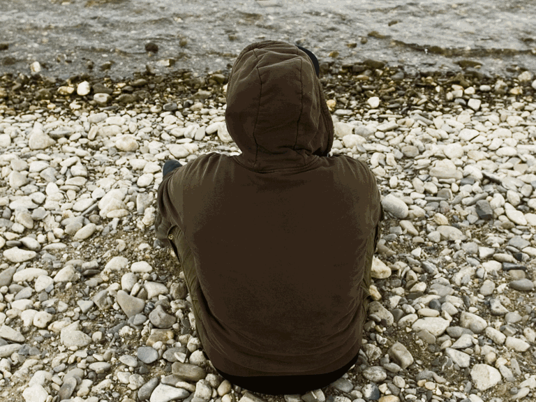 Ein junger Mann sitzt mit dem Rücken zur Kamera an einem Flußufer. Er trägt eine Shirt mit einer Kapuze.