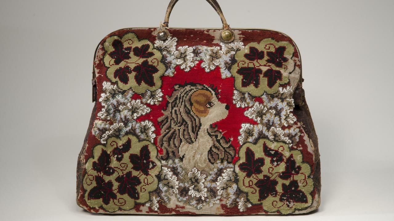 Eine Reisetasche aus Süddeutschland, etwa um 1860, aus Wollstickerei, Glas- und Metallperlenstickerei sowie Metall mit Henkel und Hundemotiv.