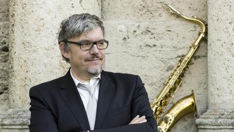 Der Jazzsaxofonist Johannes Enders steht mit verschränkten Armen lächelnd vor einer Steinwand, im Hintergrund sein Saxofon