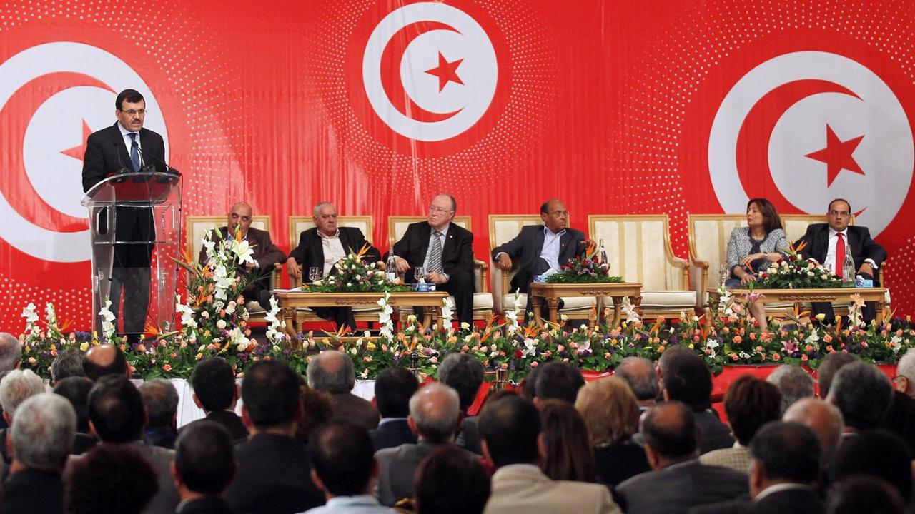 Vor einem roten Hintergrund mit drei Halbmonden sitzen die Teilnehmer des Dialogs nebeneinander auf einem Podium an einem Tisch. Der Ministerpräsident steht an einem Rednerpult und sticht in ein Mikrofon. Vor dem Podest sitzen in vielen Stuhlreihen Zuhörer.