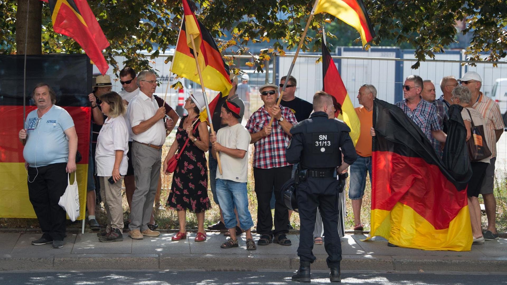Menschen stehen auf einem Bürger-Steig, mehrere von ihnen tragen Deutschland-Fahnen. Vor ihnen steht ein Polizist.