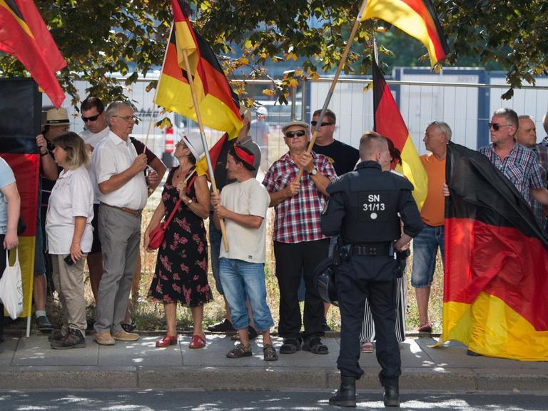 Die Demonstranten stehen auf einem Bürgersteig, mehrere von ihnen tragen Deutschland-Fahnen. Vor ihnen ein Polizist.
