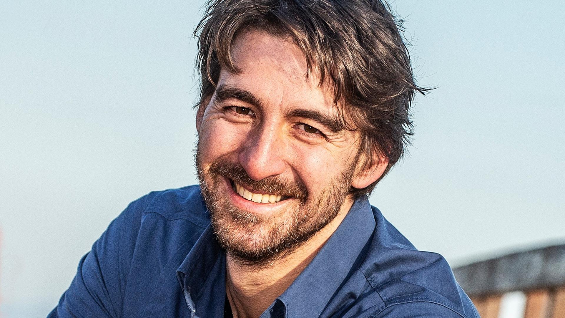 Ein Mann mit brauen Haaren und braunen Augen trägt ein dunkelblaues Hemd und lächelt in die Kamera. Im Hintergrund sind ein hellblauer Himmel und Umrisse eines hölzernen Geländers zu sehen. Ein Mann mit brauen Haaren und braunen Augen trägt ein dunkelblaues Hemd und lächelt in die Kamera. Im Hintergrund sind ein hellblauer Himmel und Umrisse eines hölzernen Geländers zu sehen.