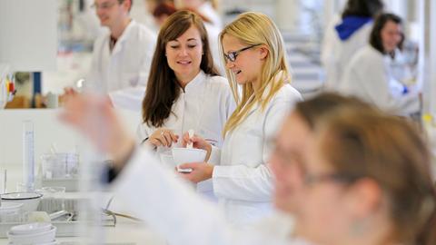 Mehrere Studenten in weißen Kitteln arbeiten in einem Labor. Eine Studentin hat einen Mörser in der Hand. Andere betrachten Reagenzgläser.