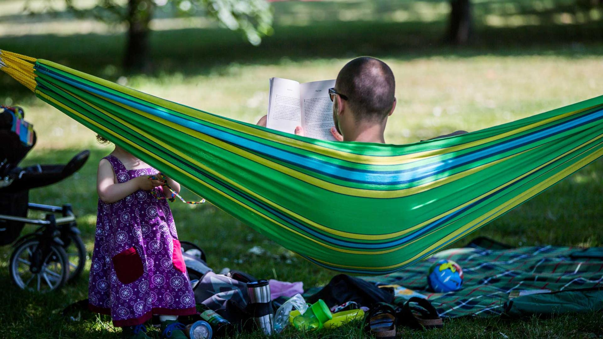 Ein Vater liest in Frankfurt am Main ein Buch in einer Hängematte im Günthersburgpark. Vor ihm spielt seine kleine Tochter im Schatten der Bäume.