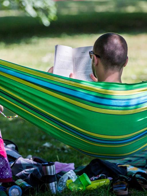 Ein Vater liest in Frankfurt am Main ein Buch in einer Hängematte im Günthersburgpark. Vor ihm spielt seine kleine Tochter im Schatten der Bäume.