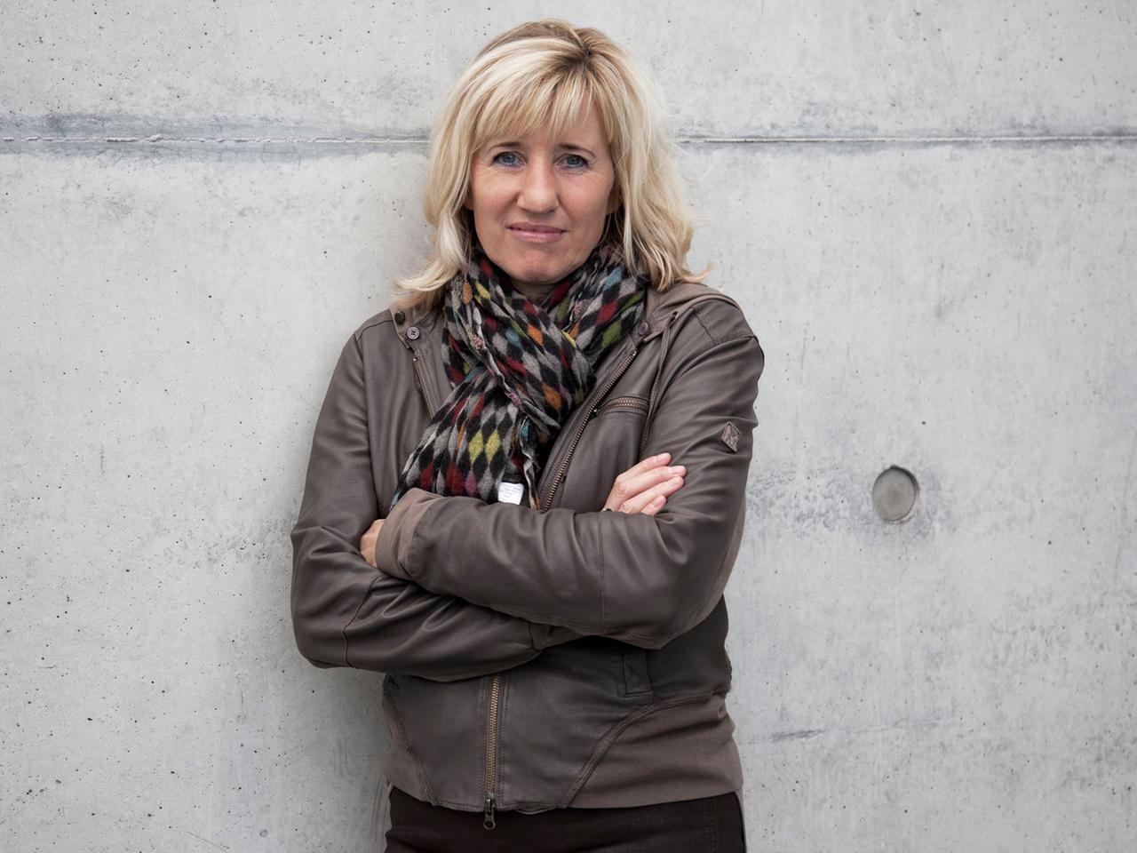 Die Schriftstellerin und Doping-Expertin Ines Geipel, die Arme verschränkt, an eine graue Betonmauer gelehnt.