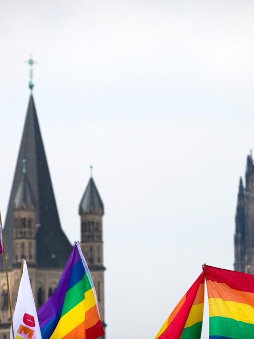 Regenbogen Fahnen im Vordergrund. Dahnter ist verschwommen der Kölner Dom zu sehen.