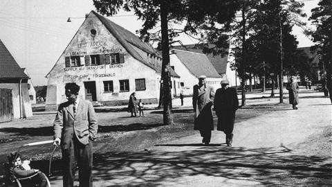 Das Schwarz-Weiß-Bild zeigt eine Straßenszene im DP-Lager Föhrenwald in Bayern mit mehreren Menschen, im Hintergrund Bäume und Gebäude.