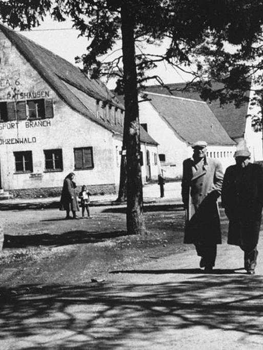 Das Schwarz-Weiß-Bild zeigt eine Straßenszene im DP-Lager Föhrenwald in Bayern mit mehreren Menschen, im Hintergrund Bäume und Gebäude.