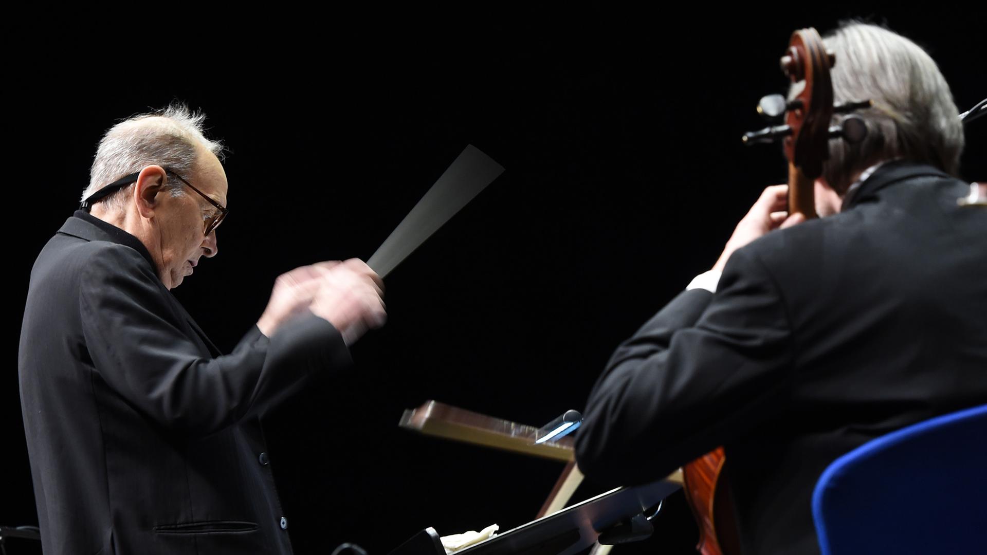 Ennio Morricone hält den Taktstock, neben ihm sitzt ein Cello-Spieler.
