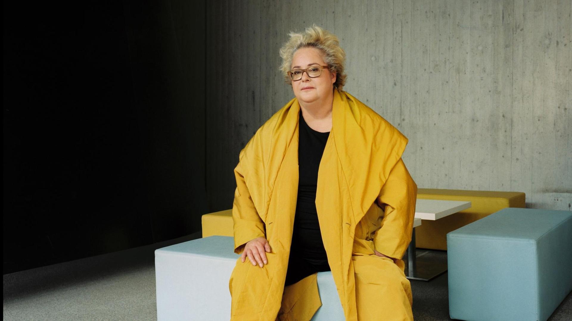 Eine Frau in weitem gelben Mantel und mit hellbrauner Brille sitzt in einem Raum aus nackten Betonwänden auf einem grauen Sitzblock und schaut in die Kamera