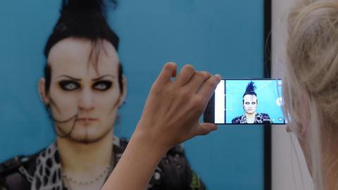 Die Arbeit "Contest 19", 2011, von Erasmus Schröter bei einer Ausstellung im Kunstmuseum Moritzburg ist auf dem Handy einer Frau zu sehen. Auf dem Foto ist ein Teilnehmer des Wave-Gotik-Festivals in Leipzig zu sehen, der sich dafür verkleidet und geschminkt hat.