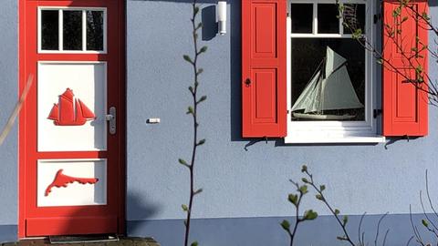 Auf dieser modernen Variante einer Darßer Tür ist die Silhouette eines Segelschiffs und ein Inselumriss zu sehen. Das Haus ist grau gestrichen, links und rechts vom Fenster sind rote Holzfensterläden.