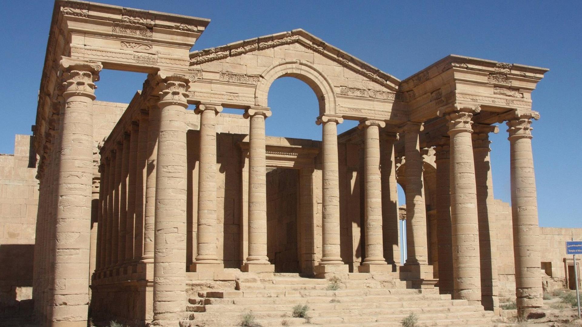 Ein antiker Tempel mit zwei Seitenflügeln und einer Treppe ohne Dach steht in der Wüste.