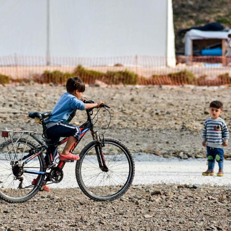 Ein Kind fährt vor Zelten auf einem für ihn zu großen Fahrrad, unweit steht ein kleineres Kind und schaut dabei zu