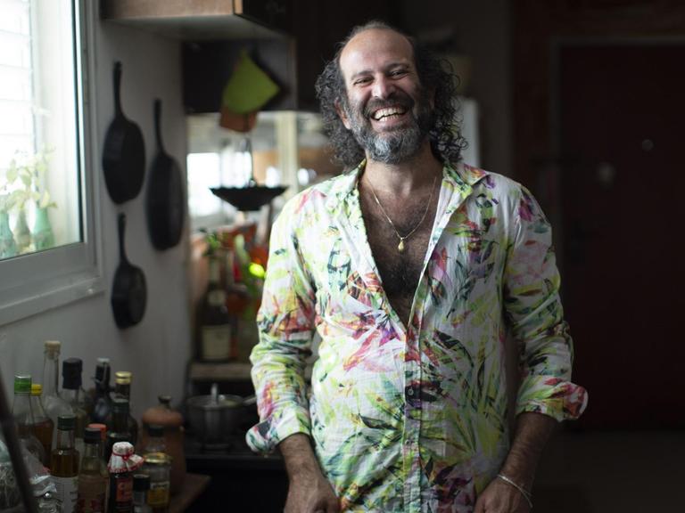 Der israelische Schriftsteller Tomer Gardi steht lachend in einem bunten Hemd in seiner Küche.